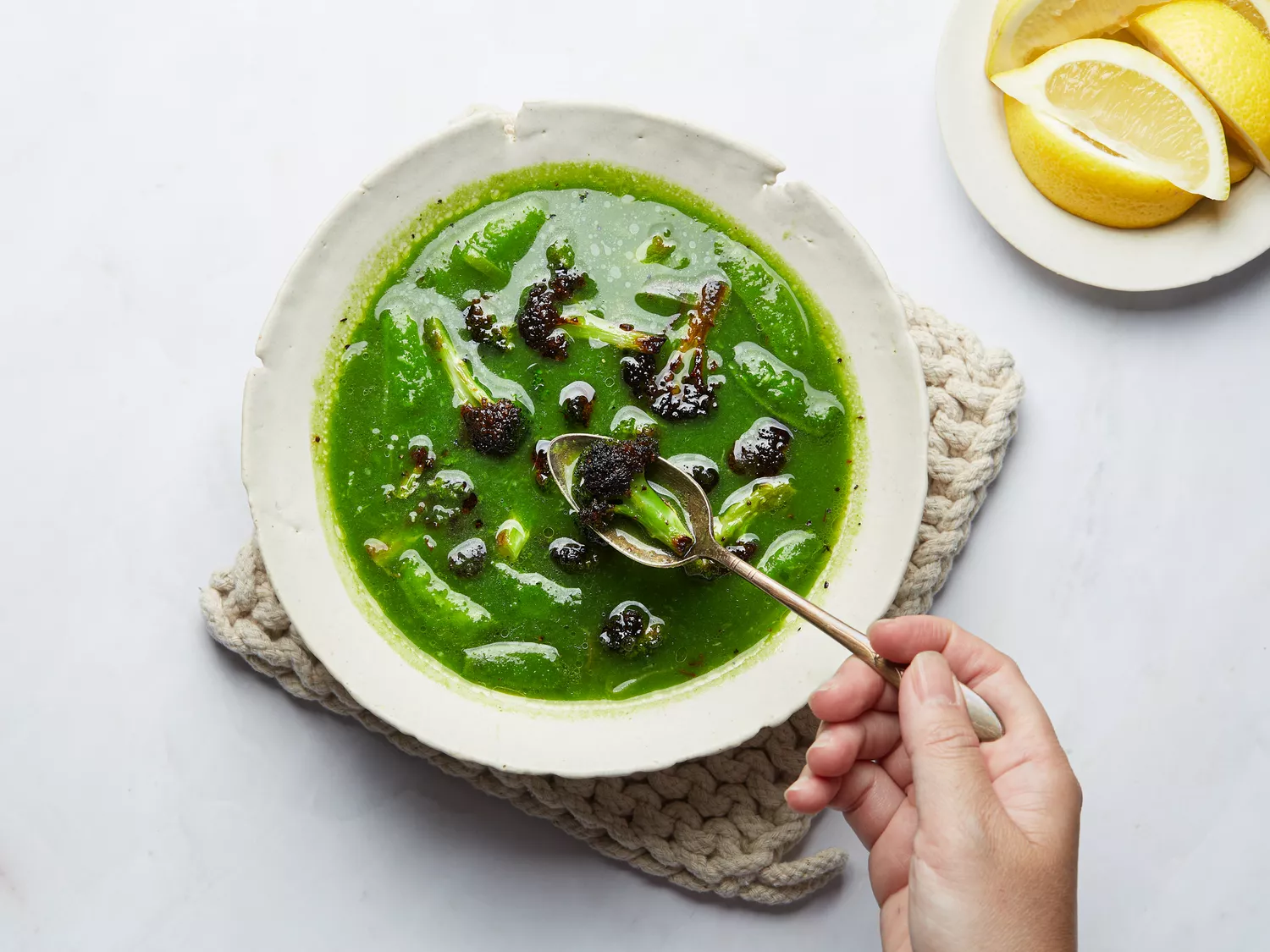 Rivoluziona la tua cucina di verdure con questo semplice trucco 7 Green Broccoli, leek and snap pea soup from above on a small woven hot pad, with a person spooning a broccoli, with lemons